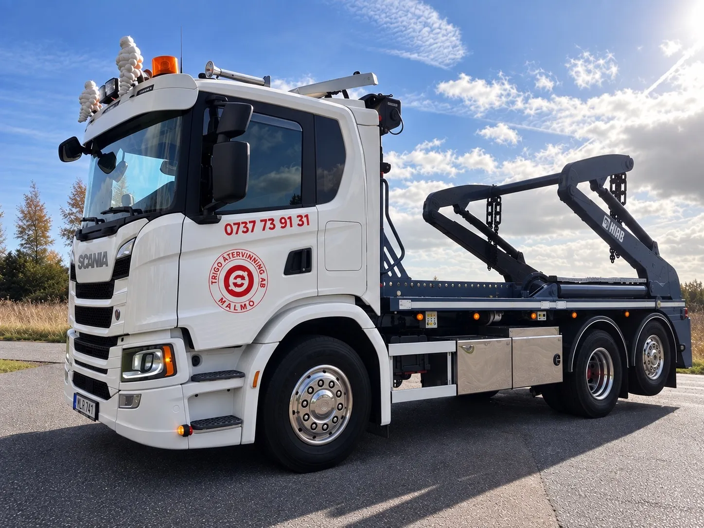 Vit liftdumperbil med lyftarmar och kedjor som lyfter en stor öppen container på en industrigård.